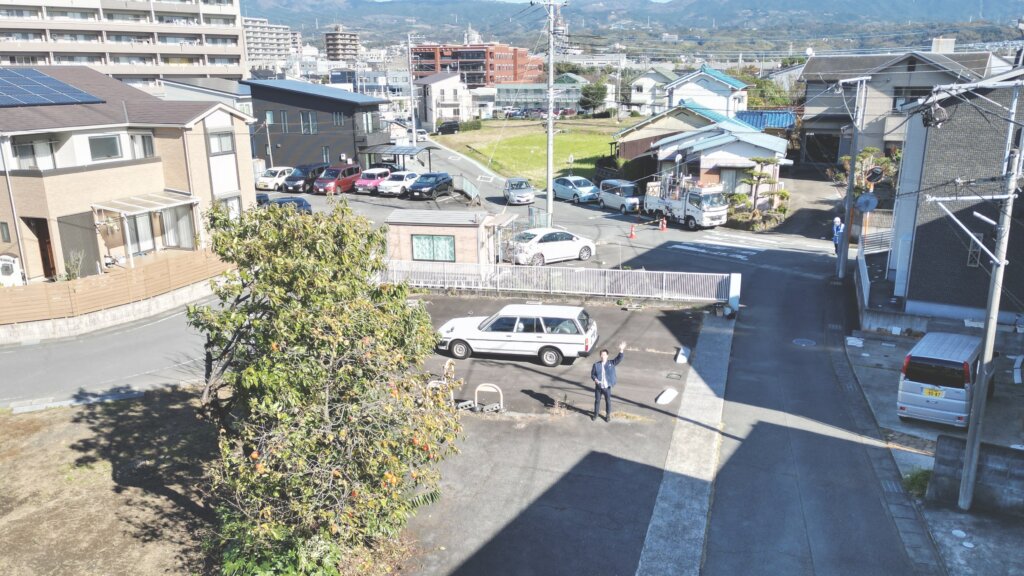 舗装された駐車場の向こうに住宅やマンションが並ぶ青木の街並みを上空から見た様子